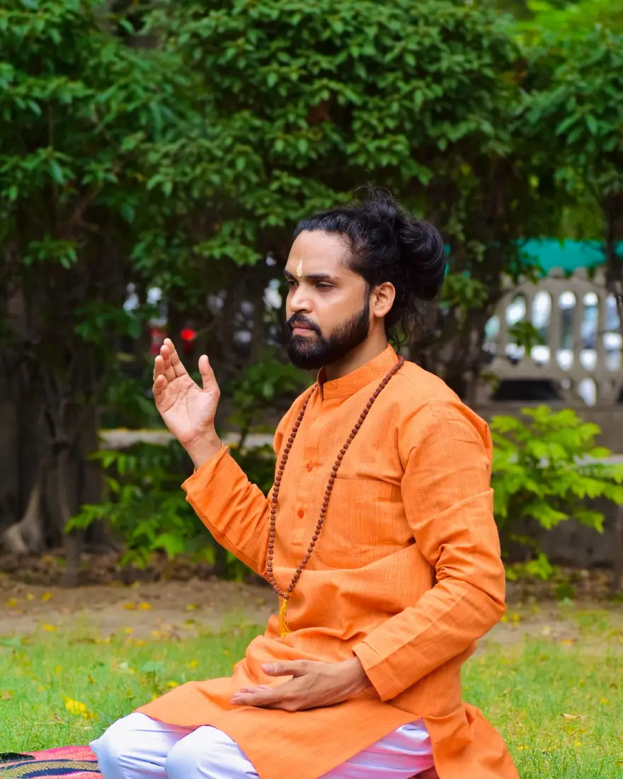YogAcharya Tarachand ji, Master Yoga Coach, seated on a mat on the grass with one palm raised as he teaches